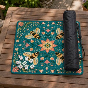 May include: A square teal mahjong mat featuring a symmetrical floral pattern with four bees surrounding a central flower, accompanied by eight white mahjong tiles and a black cylindrical carrying case with a shoulder strap.