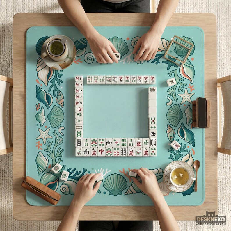 May include: A Mahjong game in play, viewed from above. The game is set on a light blue mat decorated with seashells and coral. White tiles are arranged in a square, with players' hands visible. Teacups and wooden accessories are also present.