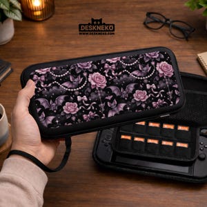 May include: Black carrying case with a floral and bat pattern. The case features a design of pink roses, bats, bows, and pearls on a black background. The case is open, revealing a black interior with storage for game cartridges.