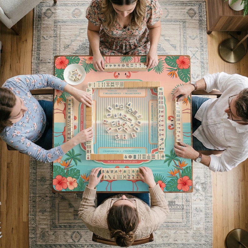 May include: A square mahjong table with a tropical flamingo and hibiscus print features a central playing area with scattered tiles, surrounded by four tile racks and a small white dish containing additional game pieces for four players.