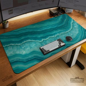 May include: A large rectangular desk mat with a teal and white marbled design. A black and gray keyboard and a black mouse sit on the mat. The mat covers a wooden desk. The brand name "DESKNEKO" is visible.