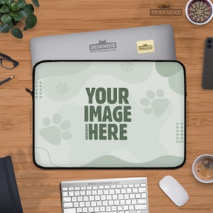 Op de afbeelding: Laptoptas met een lichtgroen pootafdrukontwerp en de tekst "YOUR IMAGE HERE". De tas ligt op een houten bureau met een laptop, toetsenbord, muis en koffiekopje. De tas heeft een zwarte rits.