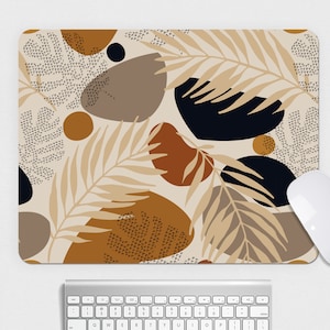 May include: A rectangular mousepad with a beige background featuring an abstract design of brown, black, and tan shapes, including leaf and dot patterns. A white computer mouse is positioned on the right side of the mousepad, with a silver keyboard below.