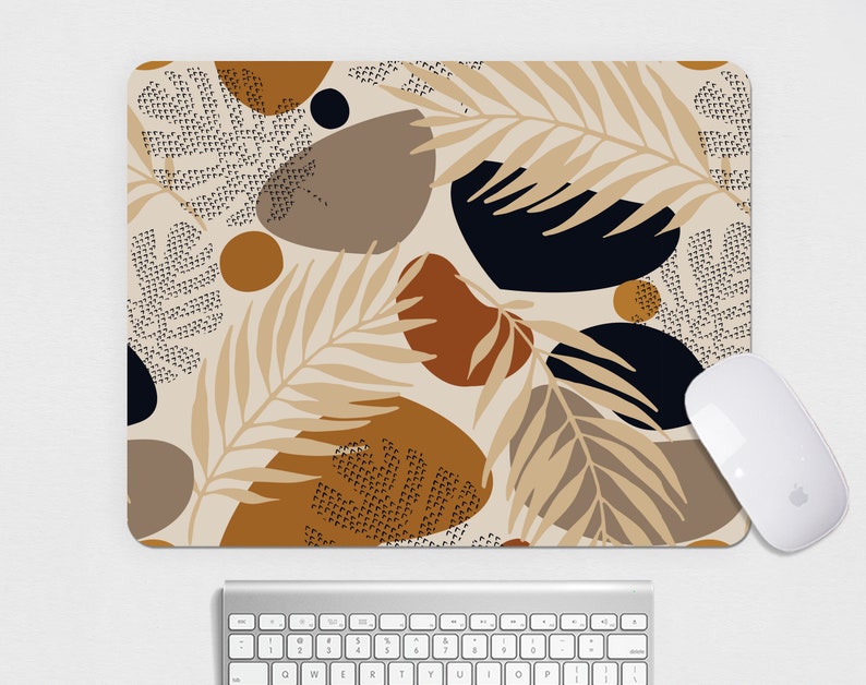 May include: A rectangular mousepad with a beige background featuring an abstract design of brown, black, and tan shapes, including leaf and dot patterns. A white computer mouse is positioned on the right side of the mousepad, with a silver keyboard below.