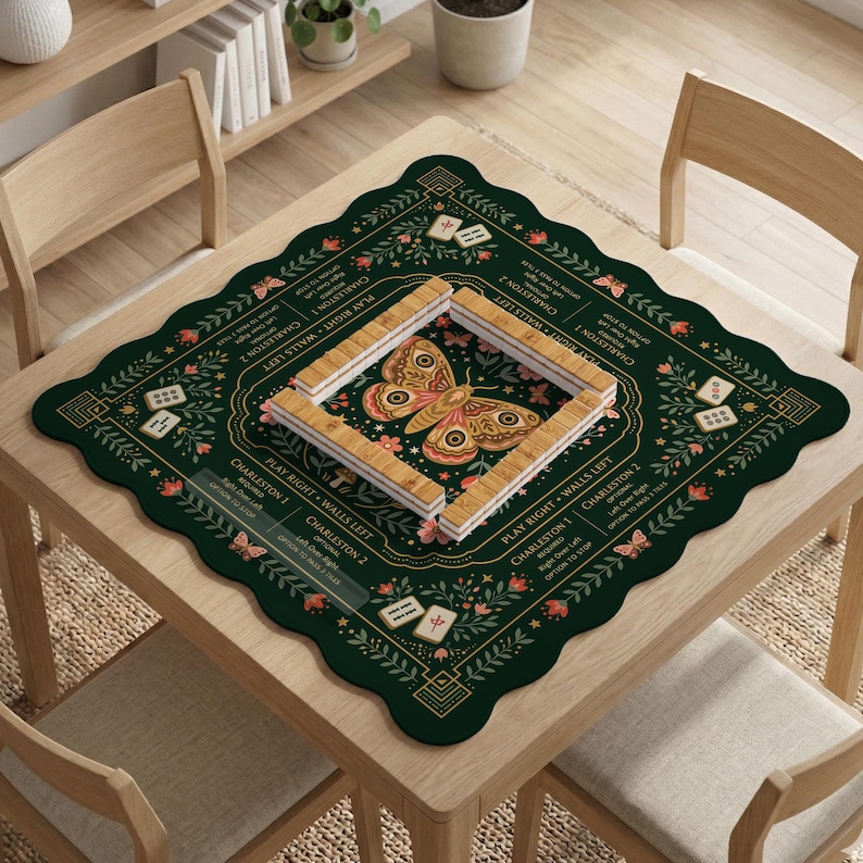 May include: A square dark green mahjong mat with scalloped edges, featuring a central moth illustration, floral patterns, and game instructions. A rectangular frame of light-colored mahjong tiles is arranged in the center of the mat.
