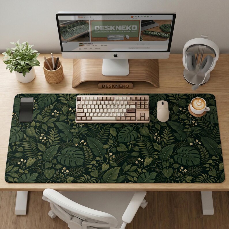 K&ouml;nnte beinhalten: Eine rechteckige Schreibtischunterlage mit einem dunkelgr&uuml;nen botanischen Muster aus verschiedenen tropischen Bl&auml;ttern und kleinen wei&szlig;en Bl&uuml;ten. Die Unterlage liegt auf einem Holztisch unter einem Computermonitor, einer Tastatur, einer Maus und einer Tasse.