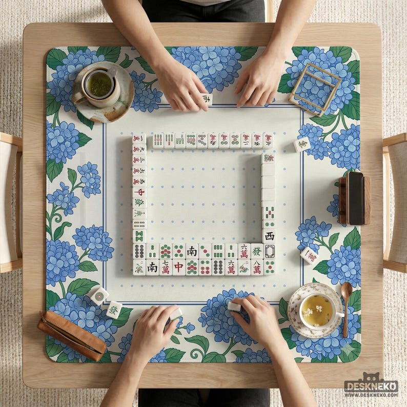 May include: Overhead shot of a Mahjong game in progress. The game is set on a wooden table with a floral patterned mat featuring blue hydrangeas. Players are arranging tiles, with cups of tea and a wooden tile holder visible.