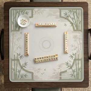 May include: A square mahjong mat featuring a light gray surface with a green bamboo and mist illustration. The mat includes four wooden tile racks holding mahjong tiles and a small white bowl containing several loose mahjong tiles.
