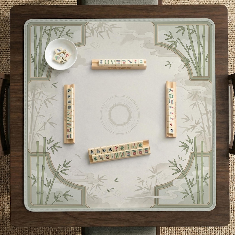 May include: A square mahjong mat featuring a light gray surface with a green bamboo and mist illustration. The mat includes four wooden tile racks holding mahjong tiles and a small white bowl containing several loose mahjong tiles.