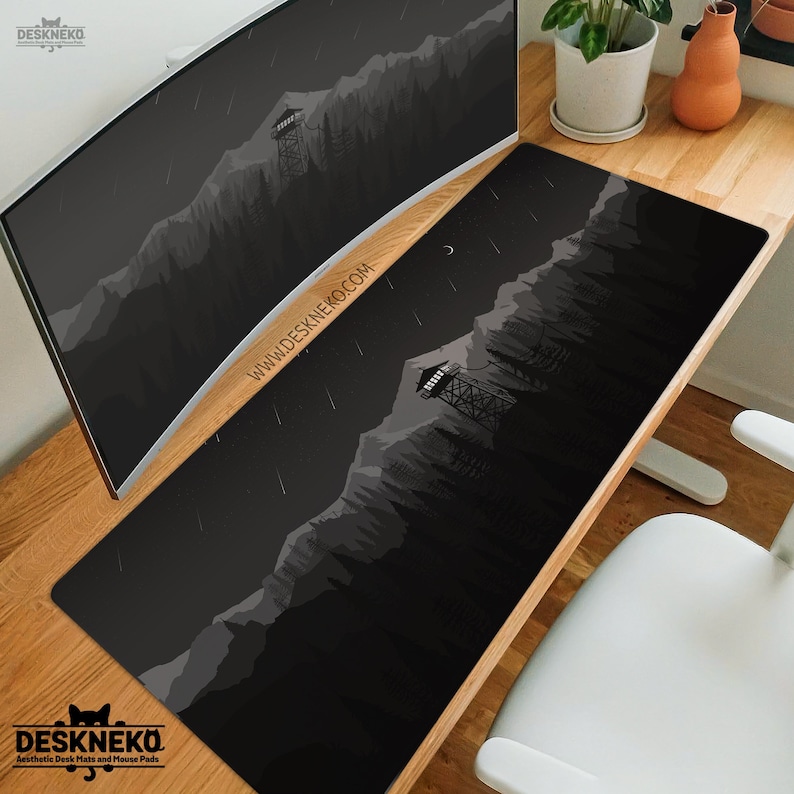 May include: Black desk mat with a minimalist mountain and forest design. The mat features a watchtower, trees, and a crescent moon. The text "WWW.DESKNEKO.COM" is printed on the mat. The DeskNeko logo is in the bottom left corner.