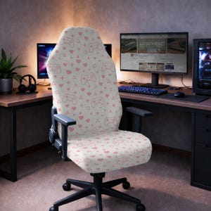 以下が含まれることがあります： ピンク色のゲームコントローラー、ハート、プラス記号のパターンが施されたクリーム色のカバーが付いたゲーミングチェア。椅子には黒いアームレストと、車輪付きの黒いベースが付いています。コンピューターモニターのあるホームオフィス環境にあります。