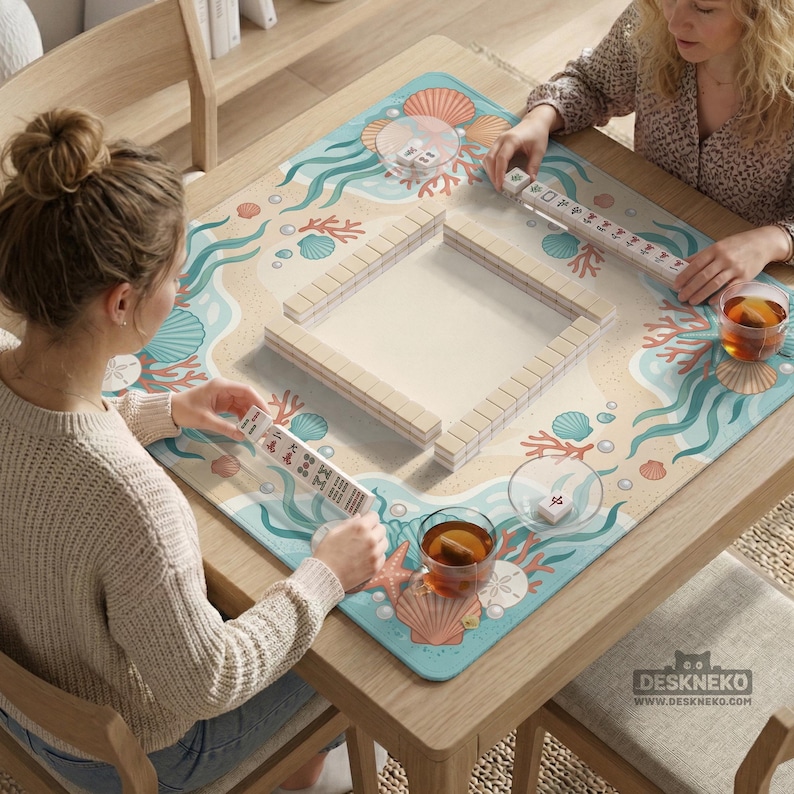 May include: A square mahjong mat featuring a beach-themed print with seashells, coral, and seaweed in shades of blue, orange, and tan. The mat displays a central square arrangement of white mahjong tiles, with additional tiles held by players.