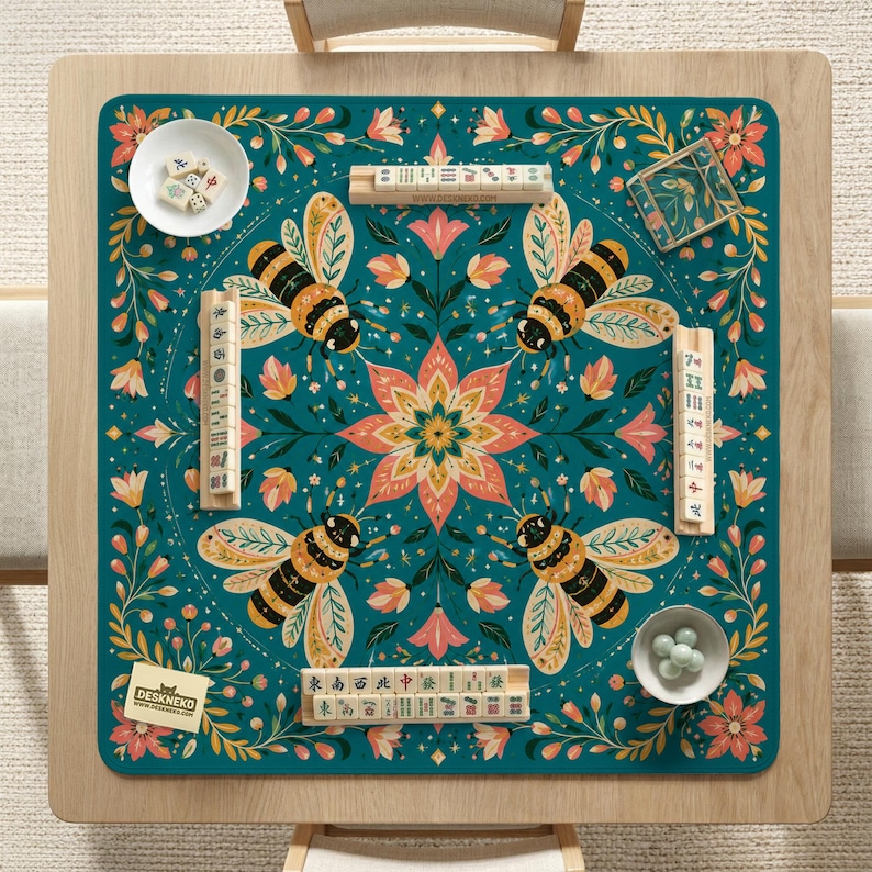 May include: A square teal mahjong mat featuring a central floral mandala surrounded by four bees and botanical patterns. The mat includes four wooden tile racks, a white bowl with mahjong tiles, a small bowl with green marbles, and a branded card.