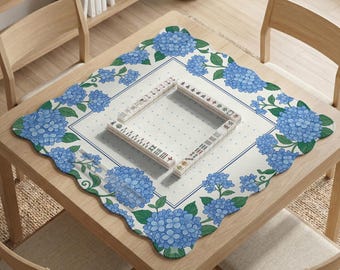 May include: A square scalloped-edge table mat featuring a white background with blue hydrangea floral patterns and green leaves. In the center, a square arrangement of white mahjong tiles with black and red characters is displayed.