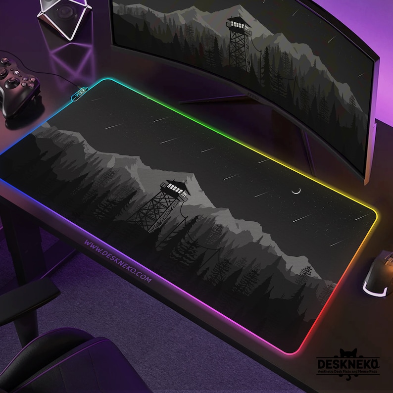 May include: A black rectangular desk mat with a mountain and forest design, a watchtower, and a crescent moon. The mat has a colorful LED border and the text "WWW.DESKNEKO.COM". A mouse and a controller are visible.