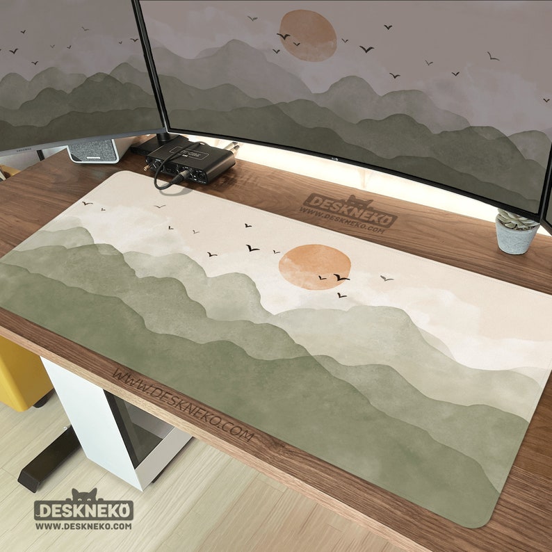 May include: A rectangular desk mat with a watercolor landscape design featuring mountains in shades of green and a setting sun. The mat has the text "DESKNEKO" and "www.DESKNEKO.COM". Black birds are flying across the sky.