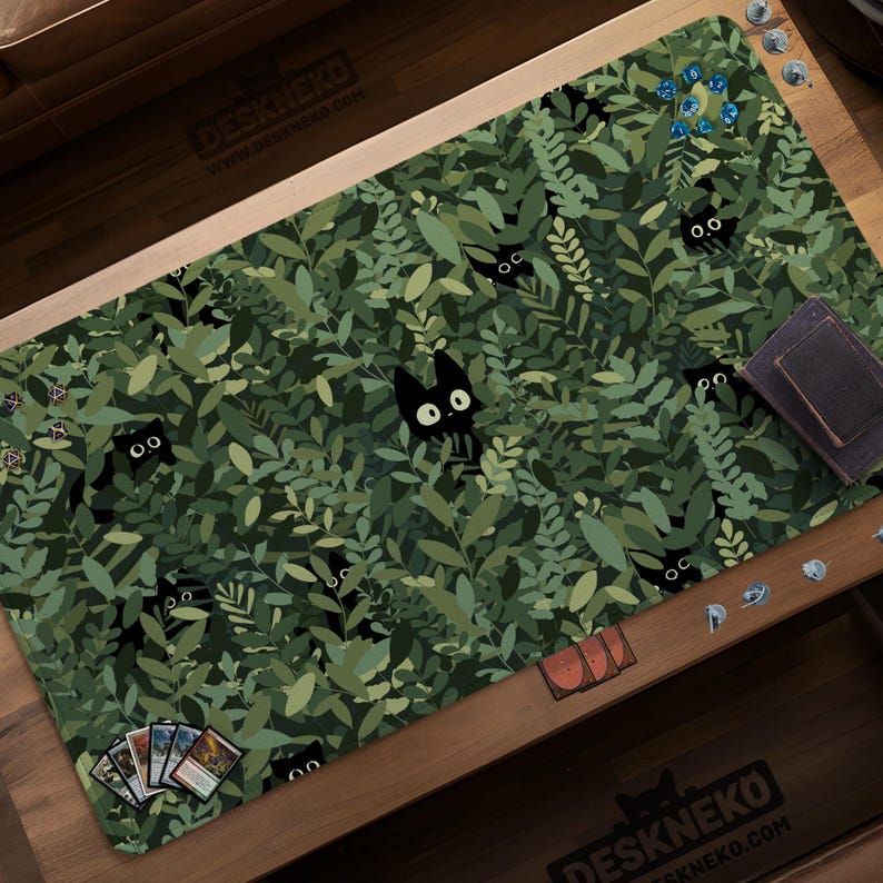May include: A rectangular play mat with a green leaf pattern and black cat illustrations. The mat has a dark border and the text "DESKNEKO" and "www.deskneko.com" printed on it. Several game accessories are on the mat.