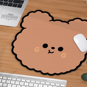 May include: A brown bear-shaped mousepad with a black border and a white mouse on a wooden desk. The mousepad has a cute cartoon face with black eyes and a small smile. The website address www.deskneko.com is visible.