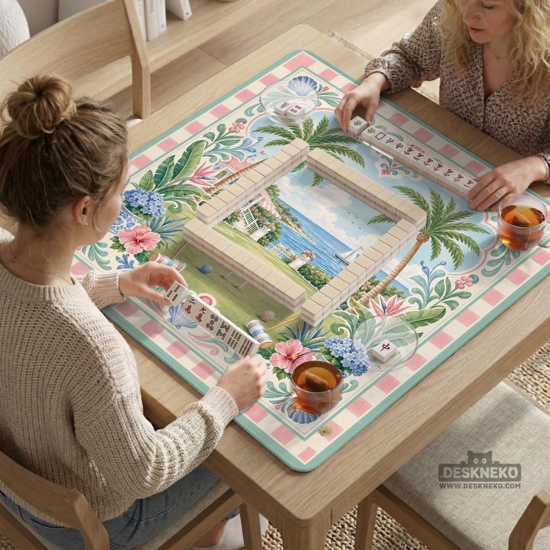 May include: Two people playing Mahjong on a wooden table. The game board features a tropical landscape design with palm trees, flowers, and a coastal scene. The table has a floral patterned mat and the text "DESKNEKO" is visible.