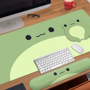 May include: A light green desk mat with a large frog design, a matching frog-shaped mousepad, and a green wrist rest. A white keyboard, mouse, and a red mug are also on the wooden desk. The text "DESKNEKO" is visible.