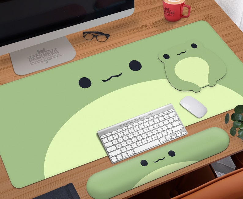 May include: A light green desk mat with a large frog design, a matching frog-shaped mousepad, and a green wrist rest. A white keyboard, mouse, and a red mug are also on the wooden desk. The text "DESKNEKO" is visible.