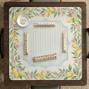 May include: A wooden mahjong table with a decorative mat featuring lemons, pink flowers, and green leaves. The mat has a light blue and white striped center. Several mahjong tiles and a small bowl with dice are arranged on the mat.
