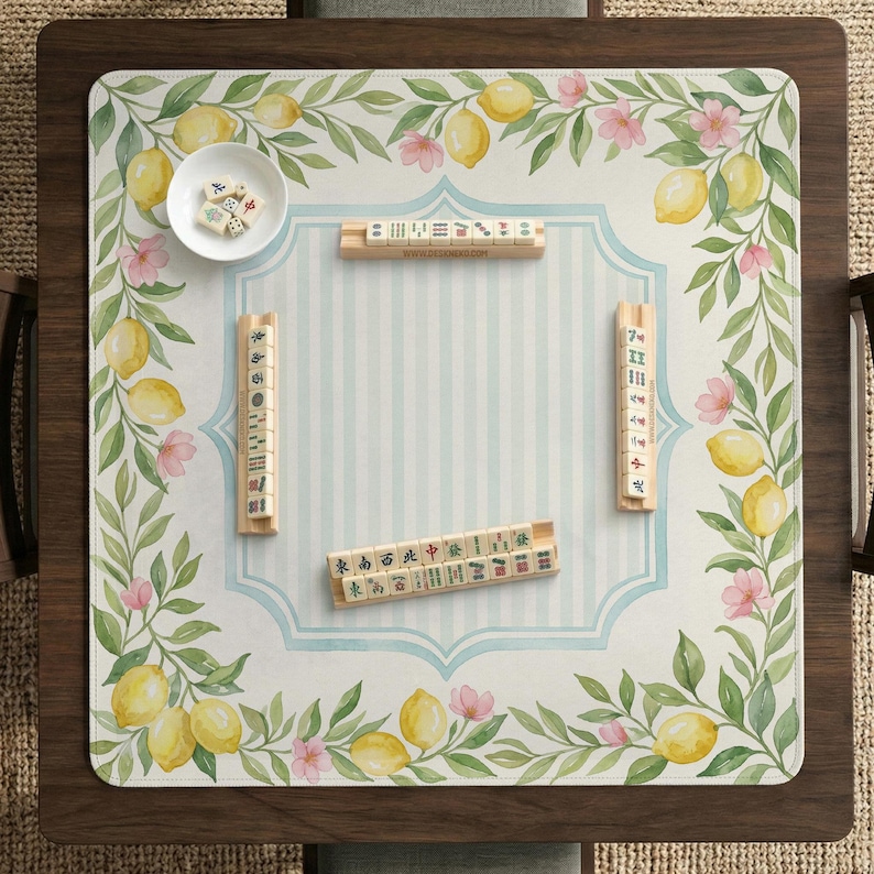 May include: A wooden mahjong table with a decorative mat featuring lemons, pink flowers, and green leaves. The mat has a light blue and white striped center. Several mahjong tiles and a small bowl with dice are arranged on the mat.
