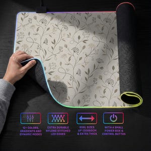 Pu&ograve; includere: Un grande tappetino per mouse rettangolare con superficie beige e un delicato motivo botanico a linee marroni. I bordi sono dotati di luci LED e il testo indica 12+ colori, bordi in nylon cuciti, dimensioni 120x60cm e una scatola di alimentazione.