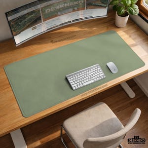 May include: A sage green desk mat with a keyboard and mouse on a wooden desk. The mat has rounded corners and the text "WWW.DESKNEKO.COM" printed on it. A curved monitor and a plant in a white pot are also visible.