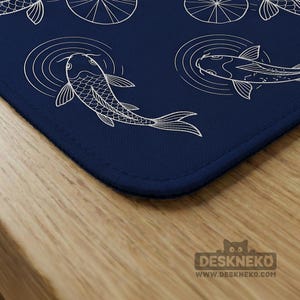 Puede incluir: Una alfombrilla de escritorio azul marino con una ilustraci&oacute;n blanca de peces koi nadando en un estanque. El dise&ntilde;o incluye c&iacute;rculos conc&eacute;ntricos para representar las ondas del agua. La alfombrilla tiene un borde cosido y est&aacute; colocada sobre una superficie de madera.