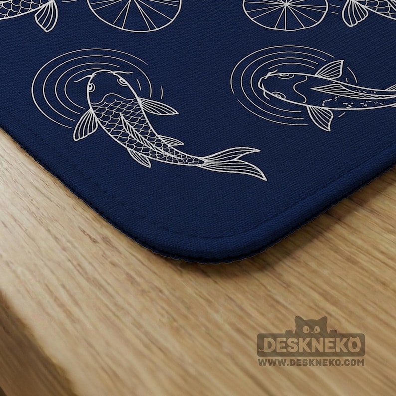 Puede incluir: Una alfombrilla de escritorio azul marino con una ilustraci&oacute;n blanca de peces koi nadando en un estanque. El dise&ntilde;o incluye c&iacute;rculos conc&eacute;ntricos para representar las ondas del agua. La alfombrilla tiene un borde cosido y est&aacute; colocada sobre una superficie de madera.