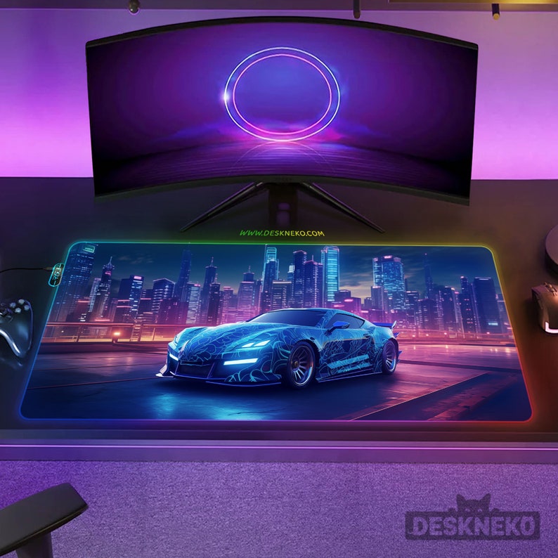 May include: A large, rectangular gaming mouse pad with a neon blue sports car design set against a cityscape backdrop. The pad has a glowing edge and the text "DESKNEKO" is visible. A curved monitor is above the pad.
