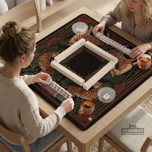May include: Two people playing Mahjong on a wooden table. The game mat features a black background with orange tigers and green foliage. The Mahjong tiles are beige, and the table has a light wood finish. Clear cups with a beverage are on the table.