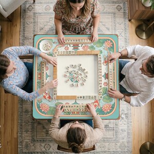 May include: Overhead view of a mahjong game in progress. A square table with a teal and floral design is surrounded by four people. The table features koi fish and lotus flowers. Wooden tiles are arranged on the table.