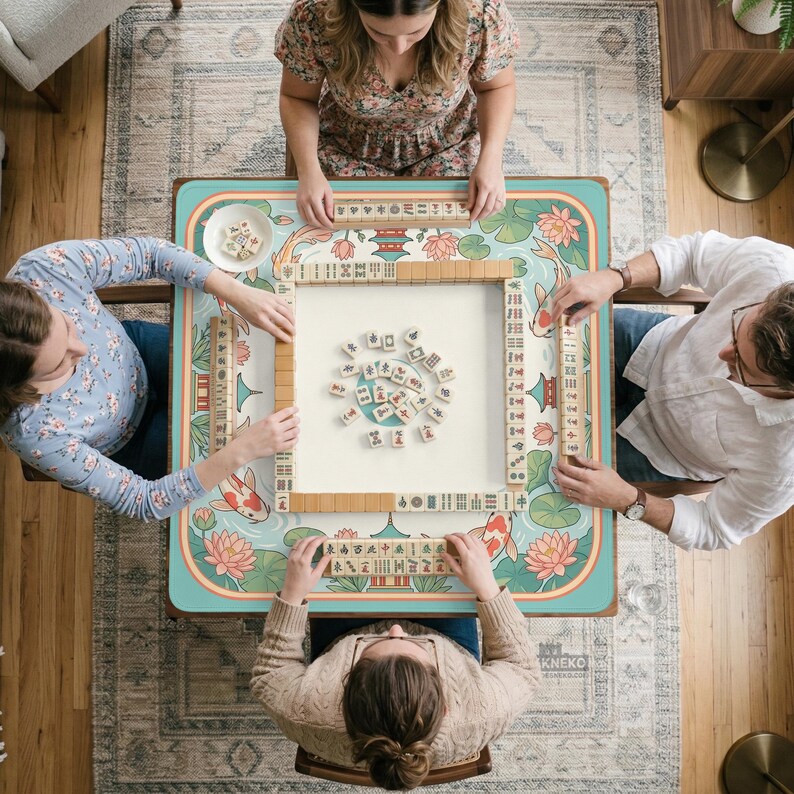 May include: Overhead view of a mahjong game in progress. A square table with a teal and floral design is surrounded by four people. The table features koi fish and lotus flowers. Wooden tiles are arranged on the table.