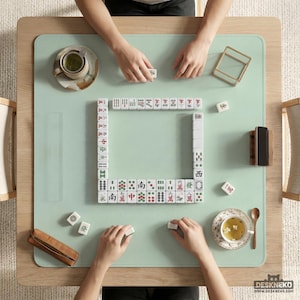 Puede incluir: Vista aérea de un juego de Mahjong en curso sobre una alfombra verde claro. La mesa de madera presenta una disposición cuadrada de fichas, con manos colocando las fichas. Tazas de té, una cuchara de madera y una pequeña caja de madera también son visibles.