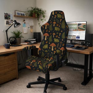 May include: A black gaming chair with a whimsical pattern of mushrooms, ferns, frogs, and keys. The chair has a high back and armrests. The design features red, brown, and green elements on a dark background. The chair is in a home office setting.
