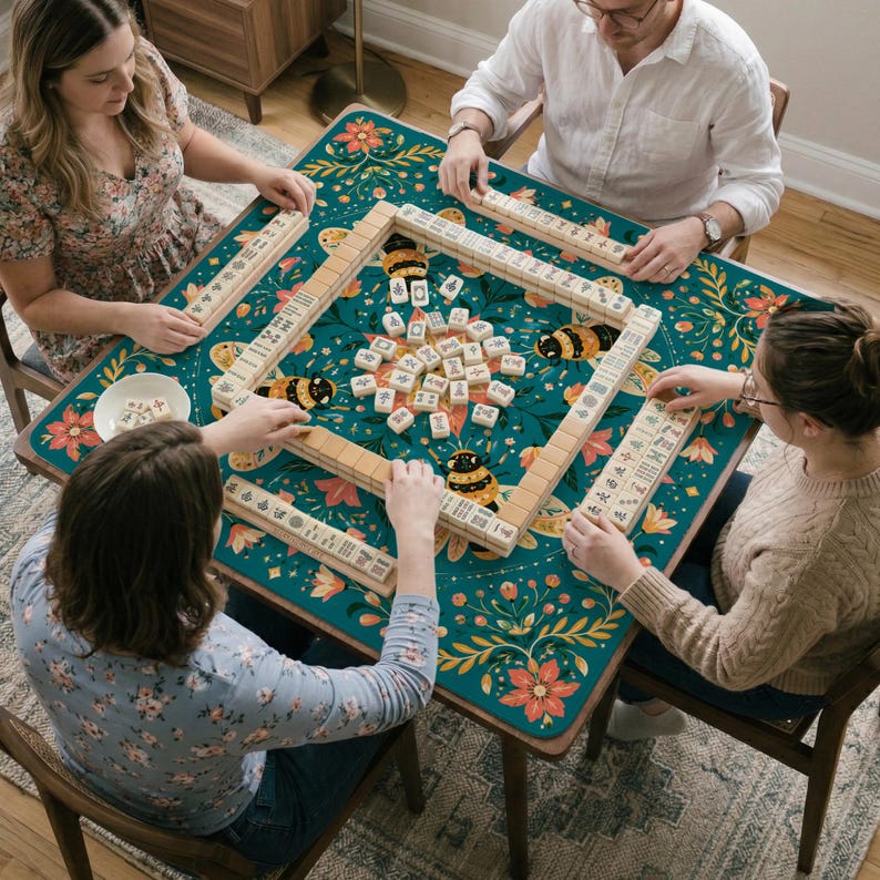 May include: A square mahjong mat featuring a dark teal background with a colorful floral pattern and bumblebee illustrations. The mat is laid out on a table with four players arranging ivory-colored mahjong tiles in a square formation.