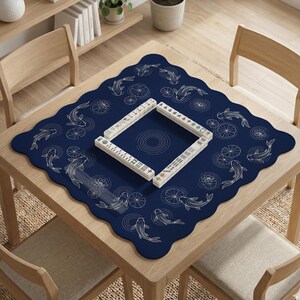 Puede incluir: Una mesa cuadrada de madera con un tapete azul marino con un dise&ntilde;o de peces koi y nen&uacute;fares. Un juego de fichas de Mahjong est&aacute; dispuesto en forma cuadrada sobre el tapete. La mesa est&aacute; rodeada de sillas de madera.