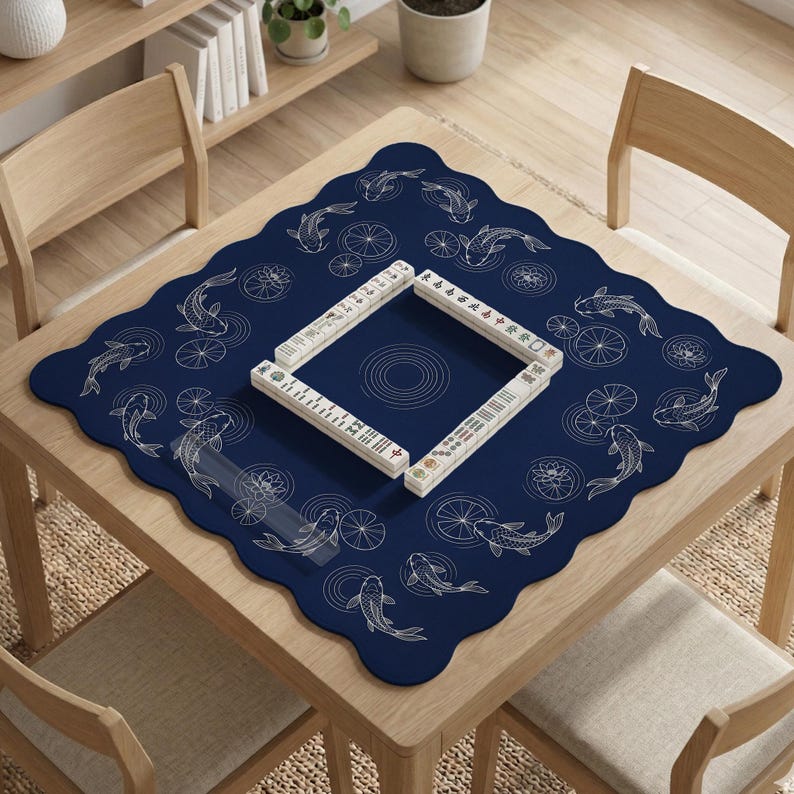Puede incluir: Una mesa cuadrada de madera con un tapete azul marino con un dise&ntilde;o de peces koi y nen&uacute;fares. Un juego de fichas de Mahjong est&aacute; dispuesto en forma cuadrada sobre el tapete. La mesa est&aacute; rodeada de sillas de madera.