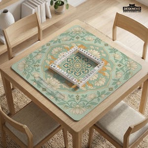 May include: A square, light green mahjong mat with a floral and dragon design, featuring a central geometric pattern. A set of white mahjong tiles is arranged on the mat. The mat is on a wooden table with chairs.