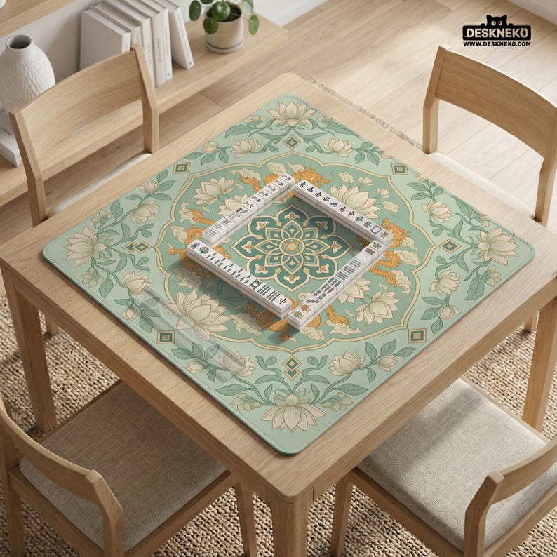 May include: A square, light green mahjong mat with a floral and dragon design, featuring a central geometric pattern. A set of white mahjong tiles is arranged on the mat. The mat is on a wooden table with chairs.