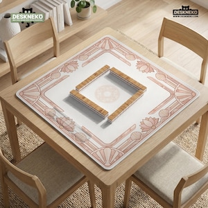 May include: A square, white mahjong mat with a pink floral and shell design. Four wooden tile holders are arranged in a square in the center. The mat is on a wooden table with four chairs. The logo "DESKNEKO" is in the top corners.