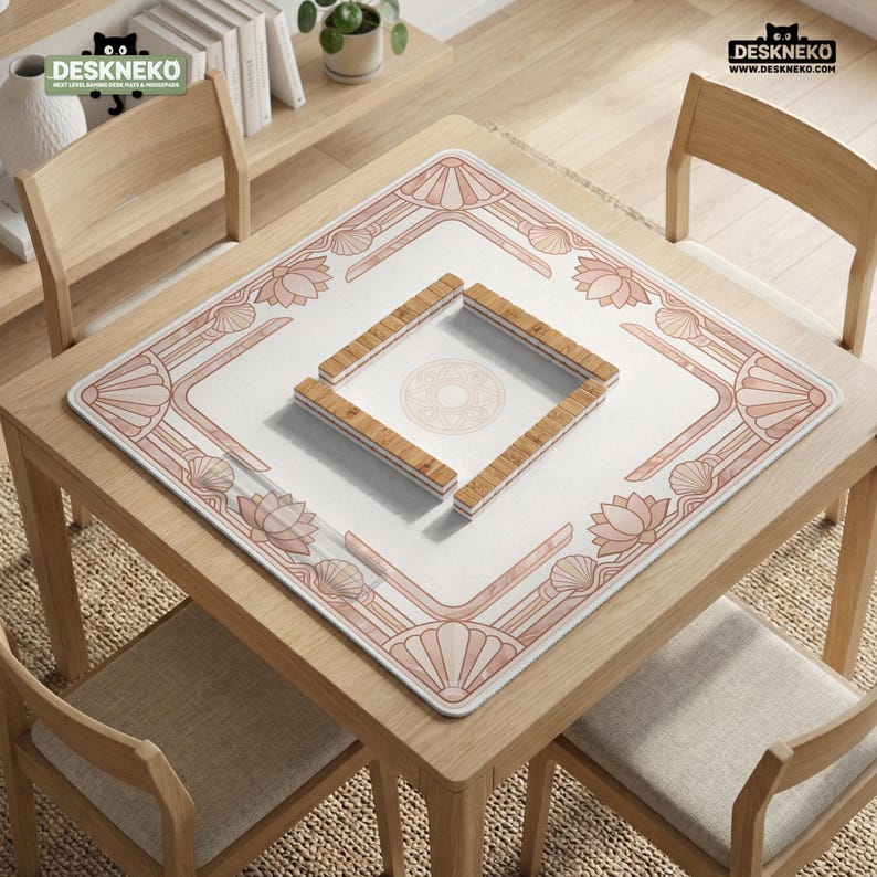 May include: A square, white mahjong mat with a pink floral and shell design. Four wooden tile holders are arranged in a square in the center. The mat is on a wooden table with four chairs. The logo "DESKNEKO" is in the top corners.