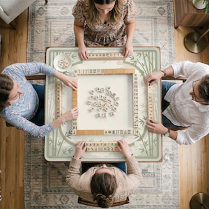 May include: A square mahjong table featuring a light green bamboo pattern border, with four players seated around it. The table surface displays a central arrangement of scattered mahjong tiles, with additional tiles organized in racks.