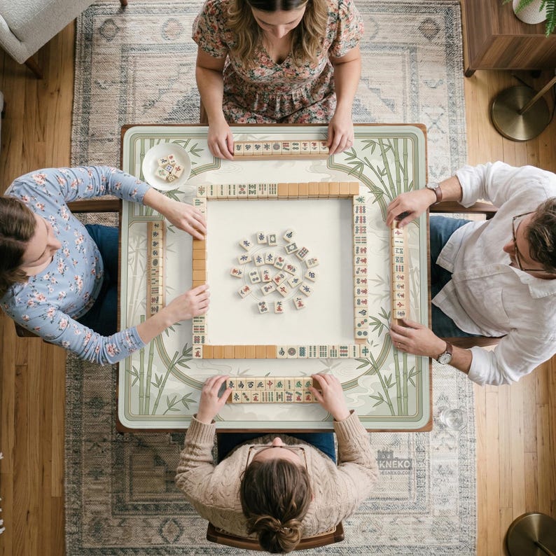 May include: A square mahjong table featuring a light green bamboo pattern border, with four players seated around it. The table surface displays a central arrangement of scattered mahjong tiles, with additional tiles organized in racks.
