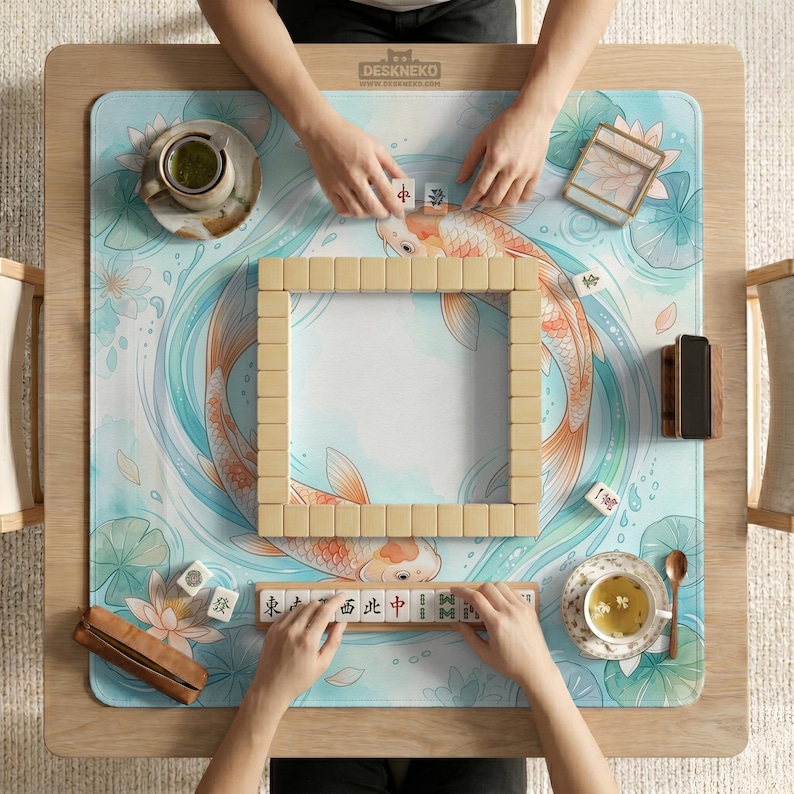 May include: A square mahjong mat featuring a light blue and white watercolor illustration of two orange koi fish swimming among lotus flowers and lily pads. The mat is set on a wooden table with mahjong tiles arranged in a square formation.