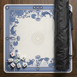 May include: A square Mahjong game mat with a blue and white floral and koi fish design. The mat has a decorative border and a central floral pattern. A black carrying case is on the right side of the mat. Several Mahjong tiles are visible.