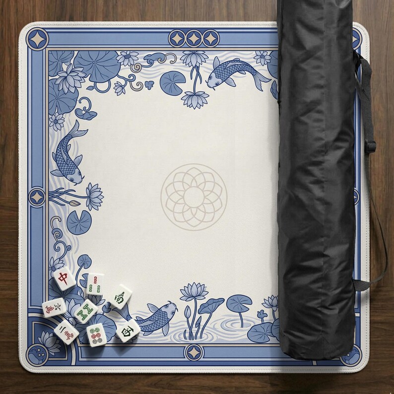 May include: A square Mahjong game mat with a blue and white floral and koi fish design. The mat has a decorative border and a central floral pattern. A black carrying case is on the right side of the mat. Several Mahjong tiles are visible.