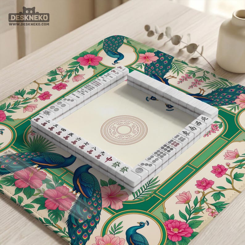 May include: A square mahjong mat featuring a cream background with a repeating pattern of blue and green peacocks, pink hibiscus flowers, and green foliage. A set of white mahjong tiles is arranged in a square frame on the mat's center.